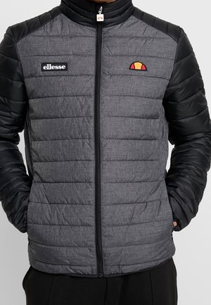 Homme portant une veste matelassée grise et noire zippée avec le logo et l'emblème ellesse sur la poitrine.