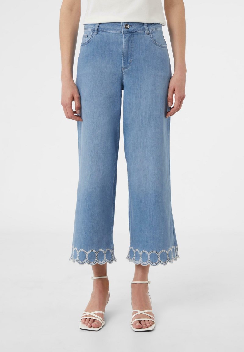 Femme portant un jean large court bleu clair avec des ourlets festonnés et brodés, des sandales blanches à brides, et un haut blanc.