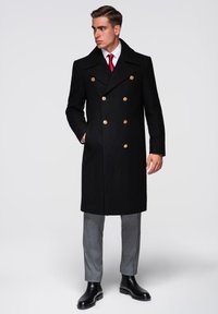 Manteau peacoat en laine noire avec un devant croisé, boutons dorés et grand col. Porté sur un pantalon gris sur mesure et des bottines en cuir noir.