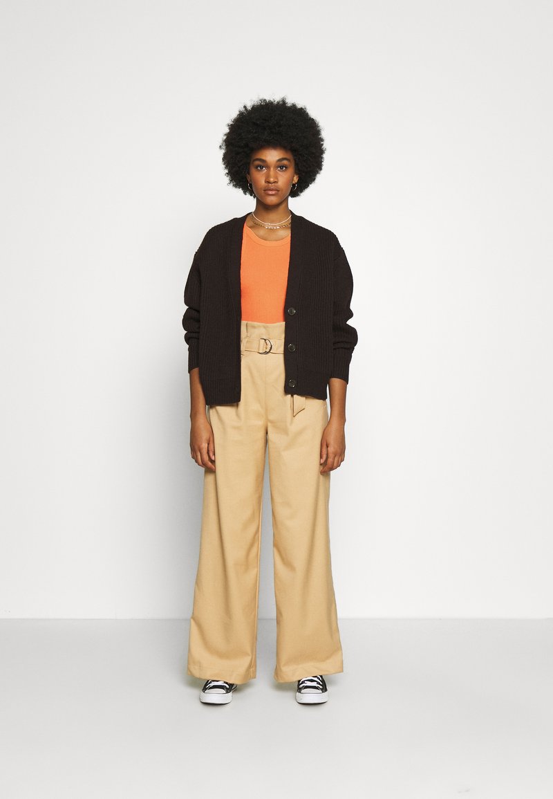 Cardigan castanho com nervuras, top laranja, calças largas bejes com cinto, sapatilhas preto e branco. Modelo em pé contra um fundo simples.