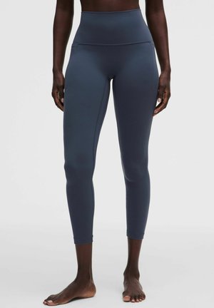 Persoon die hooggetailleerde, donkerblauwe cropped leggings draagt en blootsvoets staat op een effen lichte achtergrond.