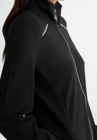 Czarna kurtka zapinana na zamek z wysokim kołnierzem, teksturowanymi panelami po bokach oraz metalicznymi zamkami na ramionach dla dodatkowego efektu.