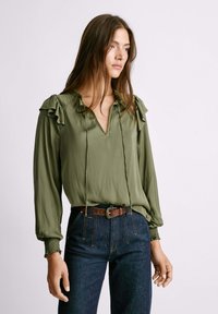 Blusa verde oliva con spalle volant, scollatura a V e dettaglio con fiocco. Tessuto liscio, maniche lunghe, abbinata a jeans blu scuro a vita alta e una cintura marrone.