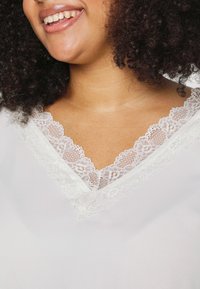 Kvinde med krøllet hår, der smiler og bærer en hvid top med V-udskæring trimmet med delikat blonde.