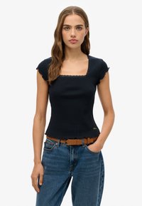 Mørkeblå ribbet top med korte ærmer, firkantet halsudskæring og blondekant. Bunden har en bølget kant. Kombineret med lyseblå jeans og et brunt bælte.