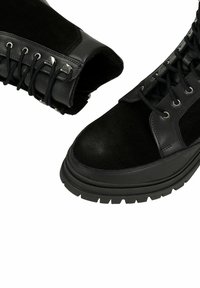 Des bottes noires à lacets montantes présentent un mélange de cuir lisse et de daim, une semelle en caoutchouc robuste et des œillets en métal. Détail de coque renforcée à l'avant.