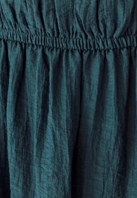 Tkanina u tirkiznoj boji sa naboranom teksturom, s elastičnom pasicom i lepršavim, nabranim dizajnom. Mekan i lagan materijal.