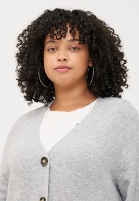 A woman with curly hair wears a gray cardigan over a white top, looking directly at the camera with a neutral expression.