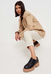Chaussures en cuir noir avec semelles épaisses et striées beige, dotées de lacets et d'une forme arrondie, associées à un blazer beige clair et un pantalon crème.