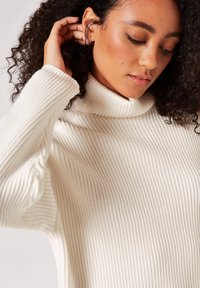 Maglione a dolcevita in crema, con una texture a coste e maniche morbide. Materiale soffice con colletto alto e vestibilità casual.