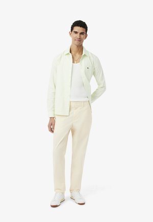 Jeune homme debout, portant une chemise verte clair boutonnée par-dessus un débardeur blanc, un pantalon crème, des baskets blanches et un collier en or.