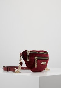 Riñonera de terciopelo rojo con cremalleras y detalles en tono dorado, que cuenta con varios bolsillos con cremallera y una correa desmontable para un uso versátil.