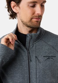 Grijze zip-up jack met een gestructureerd patroon, hoge kraag en logo op de linkerborst. Voorzien van een voorrits en een aansluitende pasvorm.