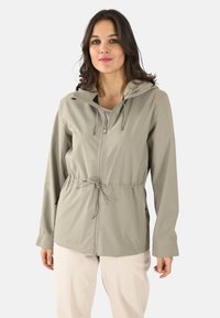 Chaqueta impermeable ligera de color beige con cremallera frontal, capucha ajustable, cinturilla elástica y bolsillos laterales. Textura suave con un diseño entallado.