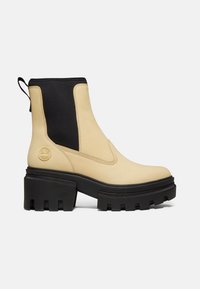 Bottine beige en cuir avec un panneau latéral élastique noir, une languette, une semelle noire épaisse à crampons et un logo rond embossé au talon.
