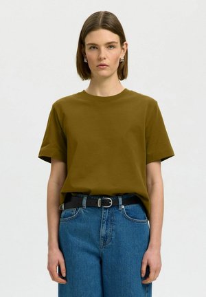 Mujer joven con el pelo corto y castaño, lleva una camiseta marrón metida en unos vaqueros azules de talle alto con un cinturón negro, de pie frente a un fondo liso.
