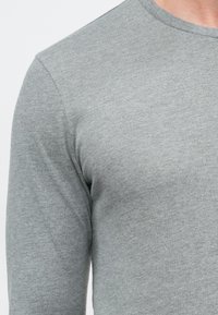 Hellgraues langärmeliges Shirt mit Rundhalsausschnitt aus einem weichen, strukturierten Stoff, der subtile Farbvariationen zeigt.