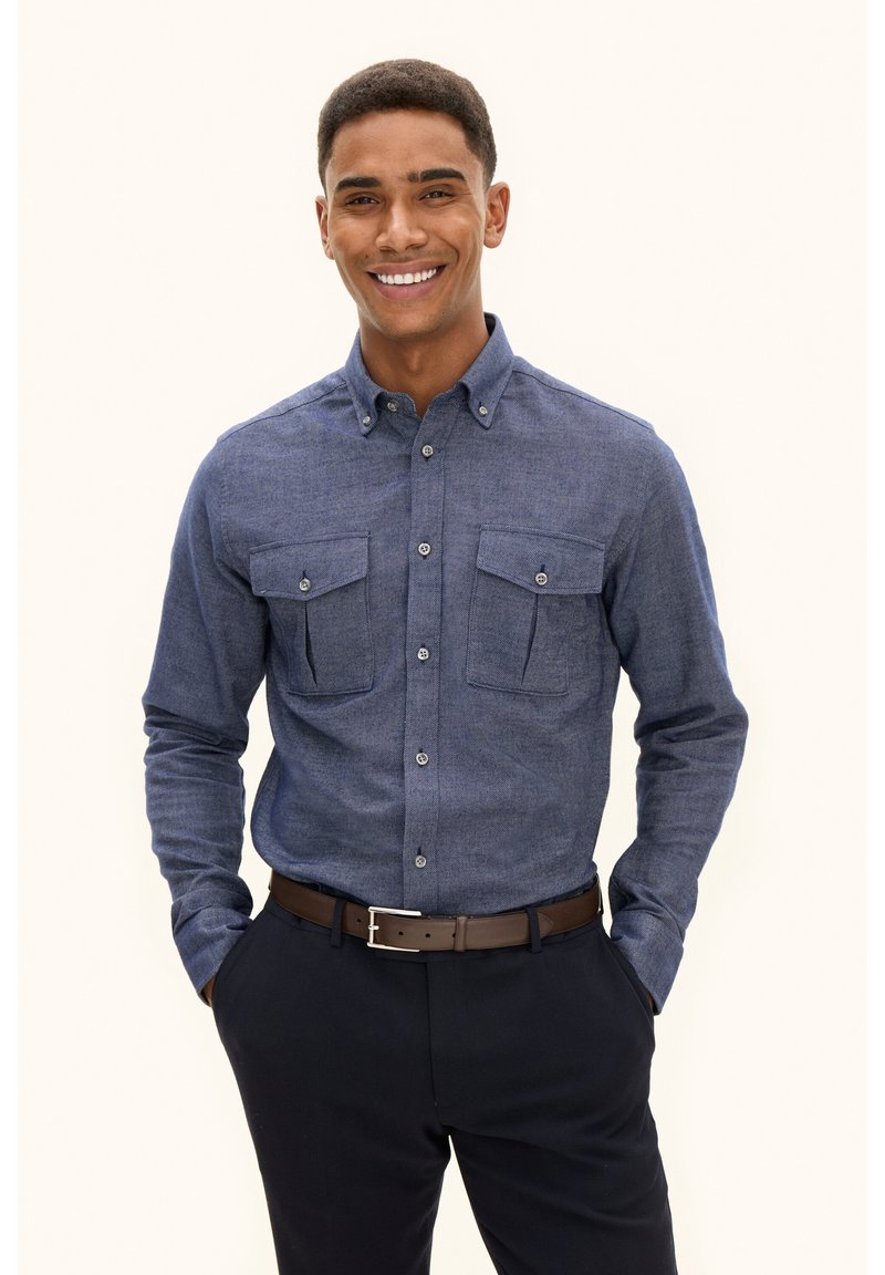 Camicia button-up blu scuro in un tessuto testurizzato, con due tasche sul petto e un colletto classico, indossata con pantaloni scuri e una cintura marrone.