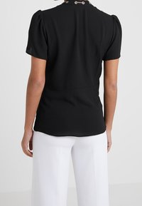 Black fitted top with short puff sleeves, elastic fabric, rounded neckline, and decorative hardware at the back. Paired with white pants.