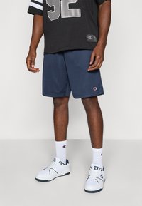 Marineblaue Netzsportshorts kombiniert mit einem schwarzen Trikot mit silbernen Akzenten und der Aufschrift "52". Weiße Sneaker und Logo-Socken runden das Outfit ab.