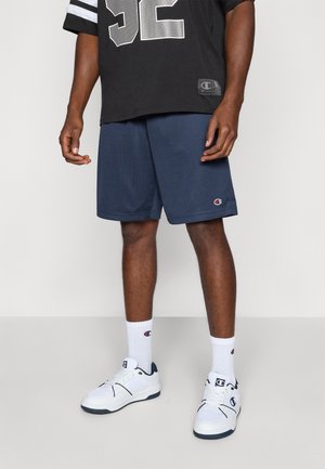 Pantaloni scurți de sport din plasă navy, asociați cu un tricou negru cu accente argintii și numărul "52" imprimat. Pantofi sport albi și șosete cu logo completează ținuta.