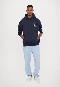 Felpa blu navy con logo bianco sul lato sinistro del petto, tasca a marsupio, cappuccio regolabile con cordino, abbinata a jeans blu chiaro e sneaker.