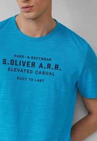 Türkise T-Shirt aus weichem Stoff, mit Rundhalsausschnitt und kurzen Ärmeln. Schwarzer Text auf der Vorderseite enthält Markenschriftzug und Phrasen.