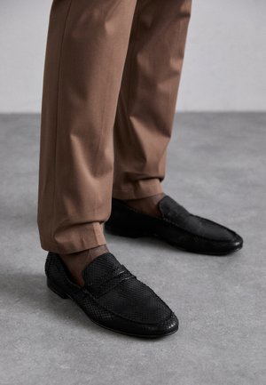 Czarne mokasyny z textured wzorem, z okrągłym noskiem i niskim profilem. Połączone z brązowymi spodniami na szarej powierzchni.