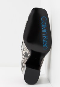 Chaussure pour femme Calvin Klein avec un motif serpent texturé noir et blanc, dotée d'un talon noir épais et d'un logo bleu distinctif sur la semelle.