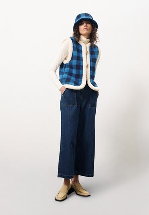 Blauw en zwart geblokte vest met crème accenten, gecombineerd met wijde denim jeans en een bijpassende bucket hat; beige loafers maken de outfit af.