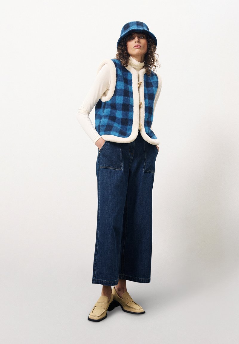 Gilet à carreaux bleu et noir avec des bordures crème, associé à un pantalon en denim à jambes larges et un chapeau seau assorti ; des mocassins beige complètent l'ensemble.