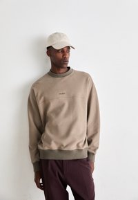 Man wearing beige sweatshirt and cap, and dark pants, standing against a plain white wall looking to the side.