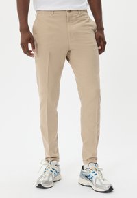 Beige Hosen mit einer taillierten Passform, die eine glatte Textur und einen einreihigen Knopfverschluss aufweisen, kombiniert mit weißen und silbernen Sportschuhen.