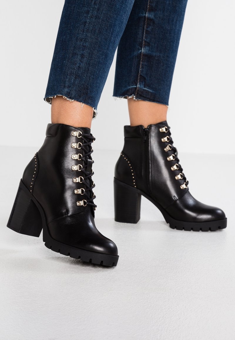 Bottines en cuir noir à talon épais, avec un laçage à l'avant, des accents dorés et des détails cloutés sur le côté.