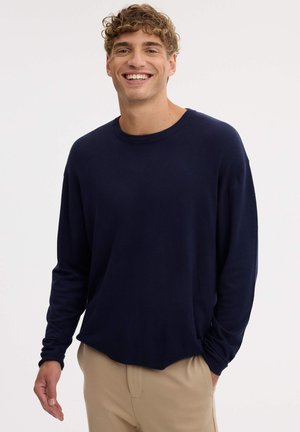 Un hombre sonriente luce un jersey azul marino de cuello redondo y unos pantalones beige sobre un fondo blanco liso.