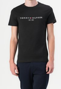 Man i svart Tommy Hilfiger t-shirt med logotyp och texten "EST. 1985", tillsammans med mörkblå byxor, står mot en vit bakgrund.