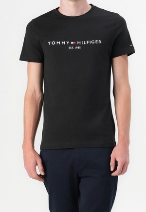 Homme portant un t-shirt noir Tommy Hilfiger avec logo et le texte "EST. 1985", associé à un pantalon bleu foncé, se tenant devant un fond blanc.