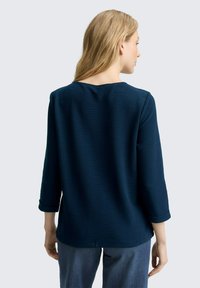 Dunkelblaues, strukturiertes Langarmoberteil mit weitem Ausschnitt und hochgerollten Ärmelbündchen, kombiniert mit hellblauen, lässig geschnittenen Jeans.