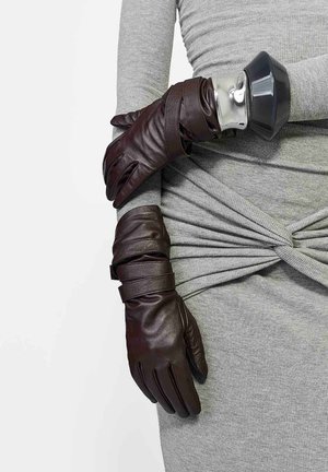 Brązowe skórzane rękawiczki z owiniętymi detalami, noszone na szarej teksturowanej odzieży, zestawione z bransoletką ze srebrnym akcentem.
