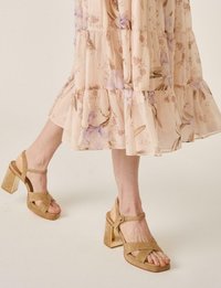 Piernas con sandalias de tacón grueso de ante beige con tiras en el tobillo, acompañadas de un vestido midi floral fluido de color rosa claro.