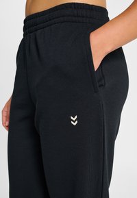 Svarta träningsbyxor med elastisk midjeresår, sidofickor och en beige chevronlogo nära midjan. Tillverkade av mjukt, hållbart tyg.