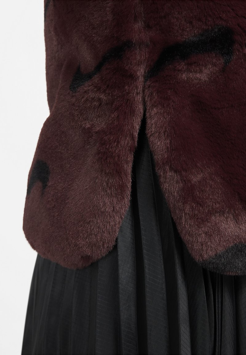 Fausse fourrure bordeaux avec une texture lisse, présentant un motif de motifs foncés, superposé sur une base en tissu plissé noir.