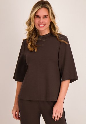 Lachende vrouw met golvend blond haar draagt een loszittende donkergroene top met ellebooglange mouwen en bijpassende broek tegen een effen achtergrond.