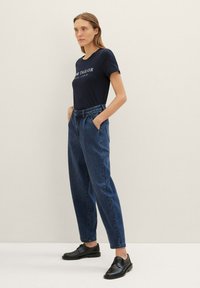 T-shirt bleu marine avec logo blanc, pantalon en denim bleu taille haute et chaussures noires. Les pantalons ont des poches latérales et une coupe fuselée.