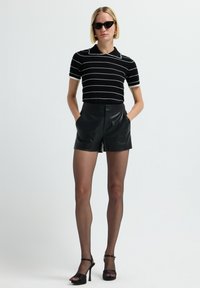 Donna con i capelli biondi corti, che indossa occhiali da sole a farfalla neri, una maglietta a righe nera a maniche corte, pantaloni di pelle neri, collant trasparenti e tacchi neri.