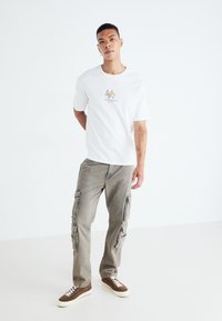 Biały bawełniany t-shirt z graficznym wzorem kwiatowym i napisem "SANTORINI", w zestawie z jasnoszarymi spodniami cargo z bocznymi kieszeniami i zamkami. Brązowe sneakersy.