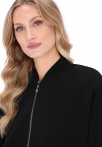 Cazadora bomber negra con cierre de cremallera y cuello acanalado; la tela tiene una textura sutil; modelo con cabello ondulado mirando a la cámara.