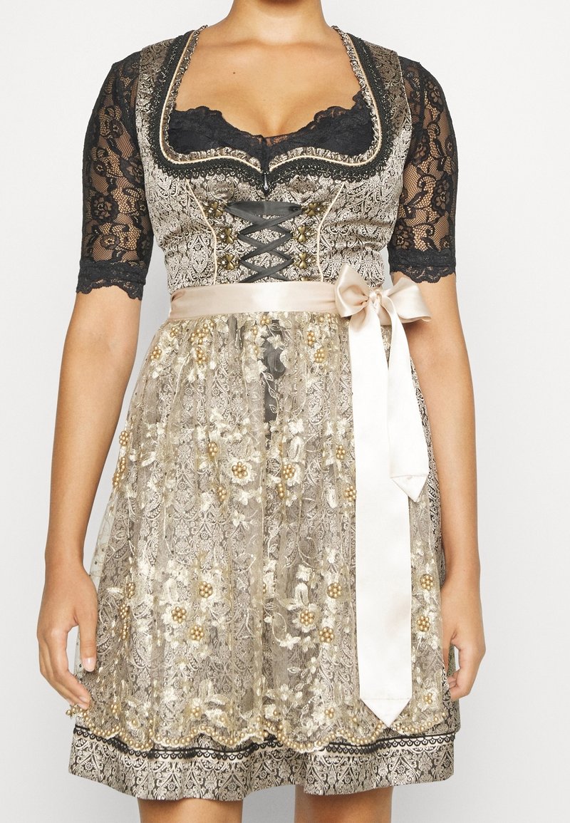 Femme portant une robe dirndl en dentelle dorée et noire avec une broderie florale et un nœud en satin attaché à la taille.