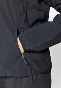 Black lightweight jacket with a textured finish, featuring an elastic cuff and side pocket with a zipper. Designed for comfort and functionality.