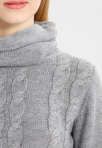 Suéter gris de punto con cuello barco, que presenta un patrón de cable texturizado a lo largo de los hombros y las mangas. Hilo suave y grueso.